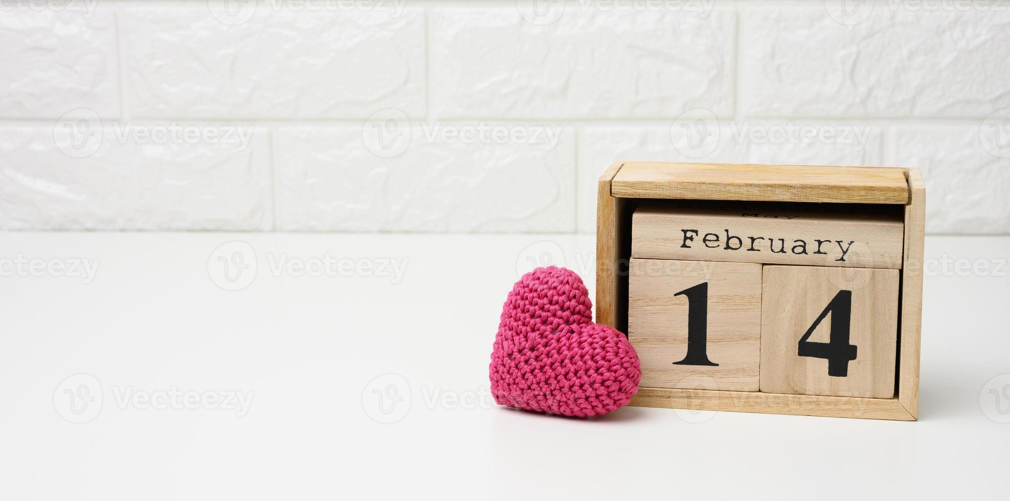 wooden calendar with date February 14 and red knitted heart, white table photo