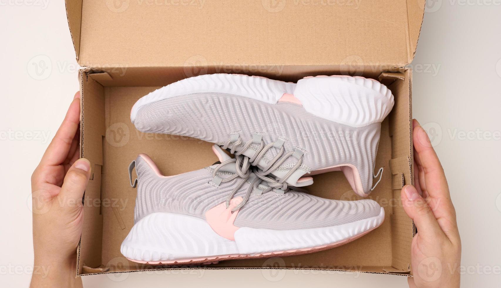 female hands are holding a brown cardboard box with gray sports sneakers on a white table, inspecting an online order photo