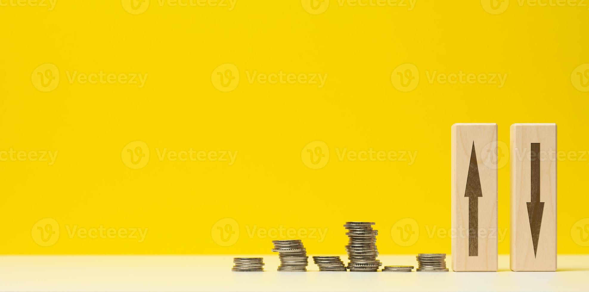 stacks of metal money and two wooden bars with arrows on a white table, yellow background. The concept of depreciation and depreciation of the exchange rate, the fall of the economy photo