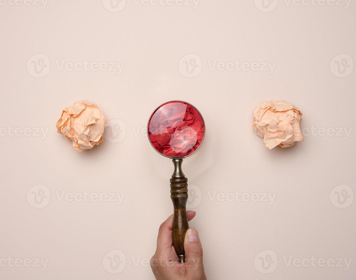 Female hand holds a wooden magnifying glass and crumpled balls of paper on a beige background photo