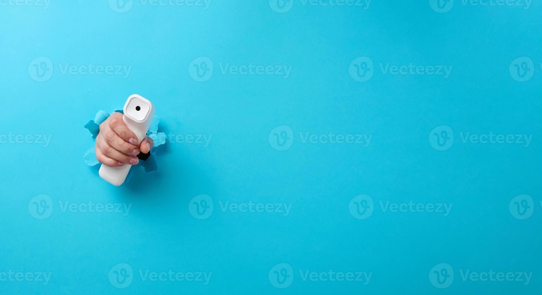 a female hand holds a plastic non-contact thermometer to measure a person's temperature, a part of the body sticks out of a torn hole in a blue paper background photo
