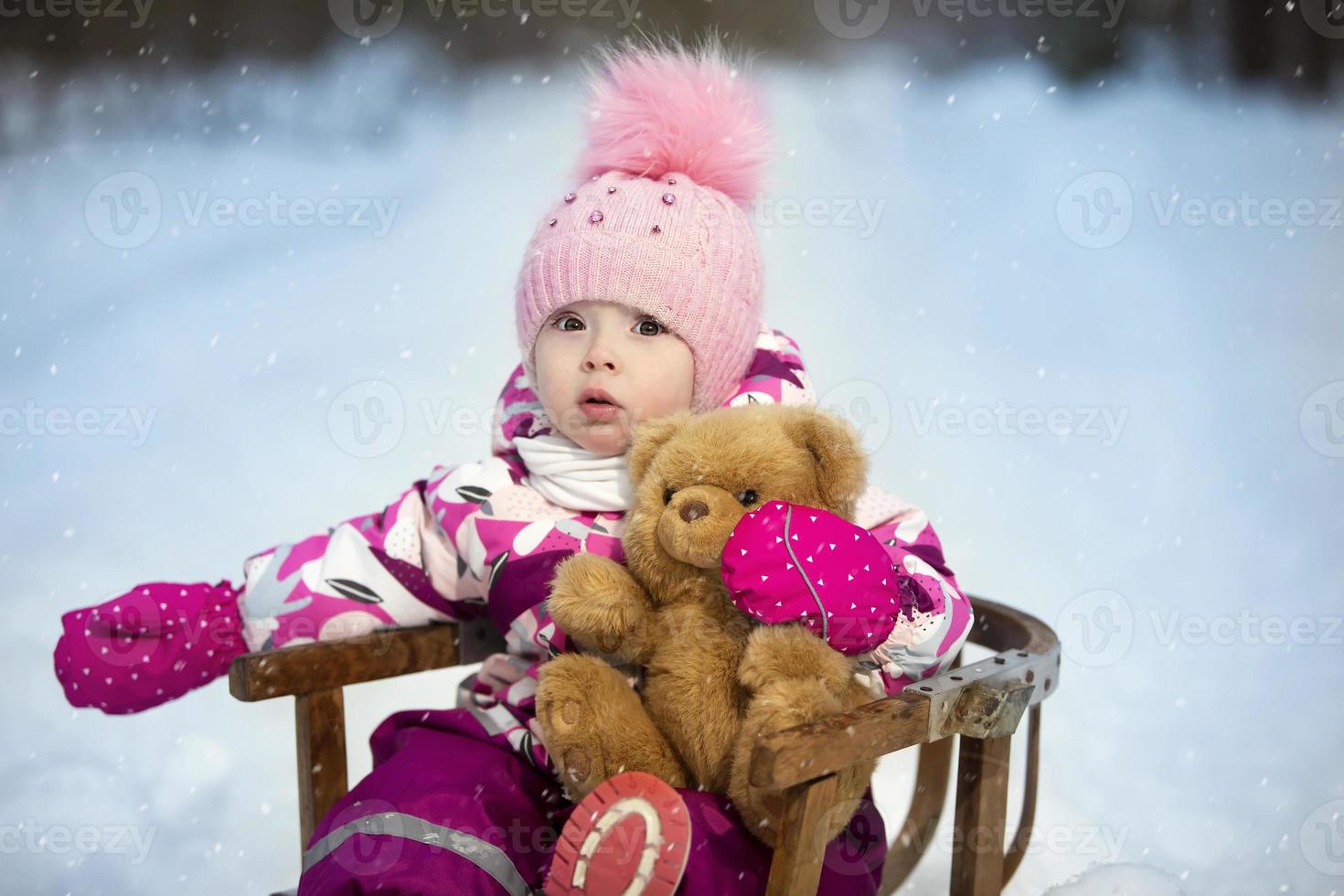 A little girl sits in a wooden sled with a teddy bear in winter. Child on a winter walk. photo