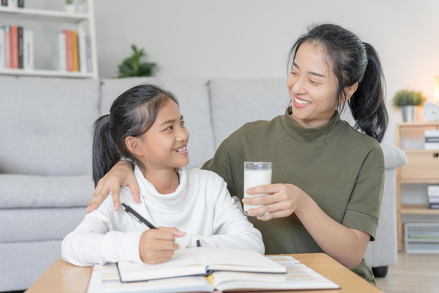 Asian young little girl learn at home. Do homework with kind mother help, encourage for exam. Mom pass on a glass of milk to daughter. Girl happy Homeschool. Mom teach and advice education together. photo