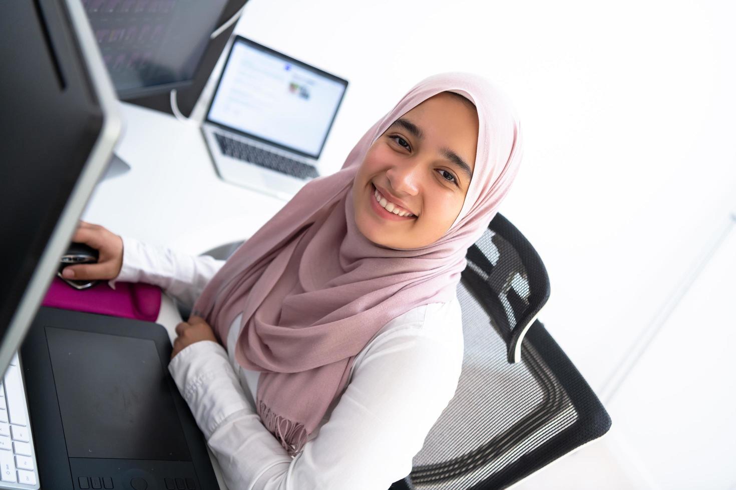 Female Arabic creative professional working at home office on desktop computer with dual screen monitor top view. Selectve focus photo