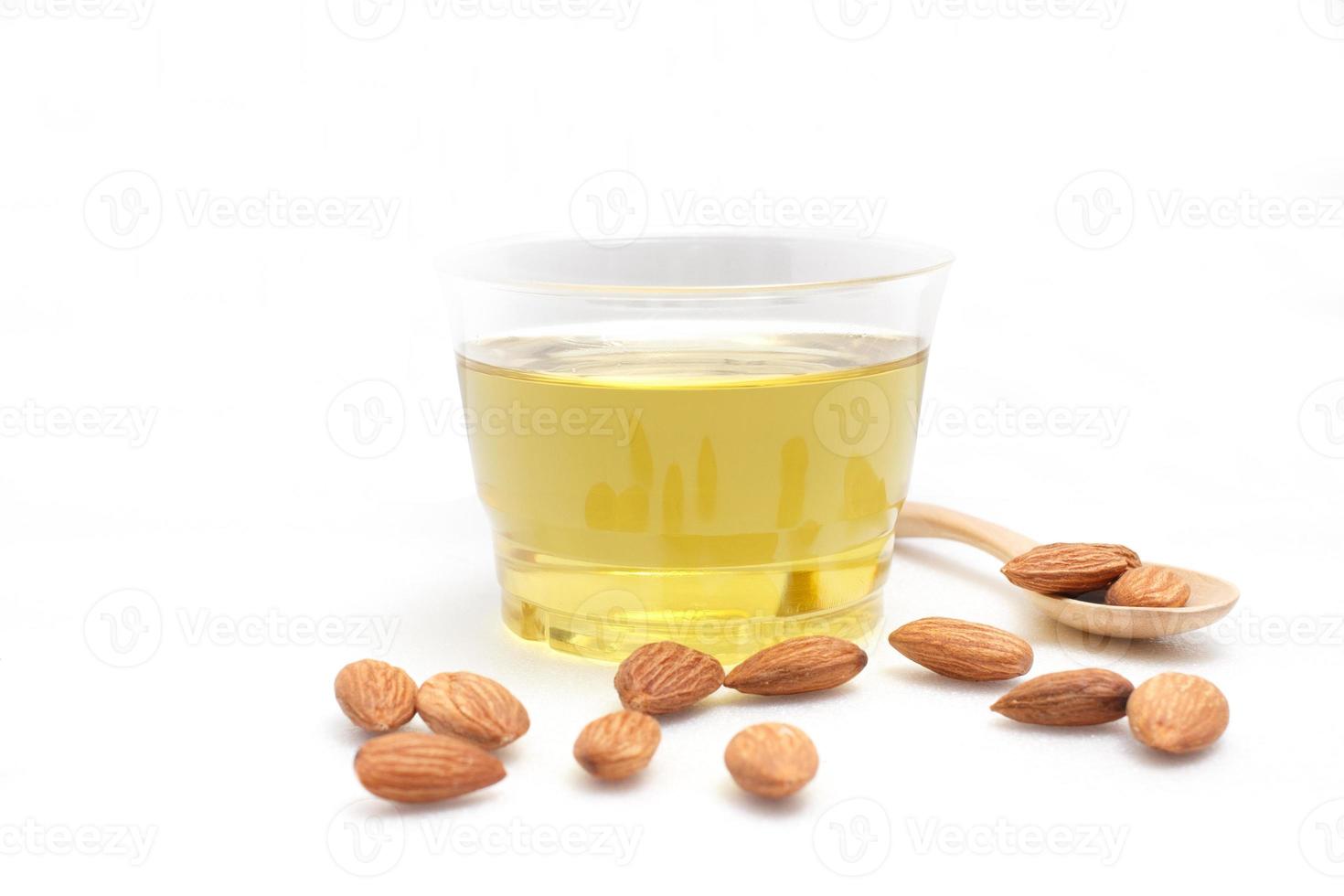 Yellow almond oil in a clear bowl with a wooden spoon next to it and several almonds on a white background photo