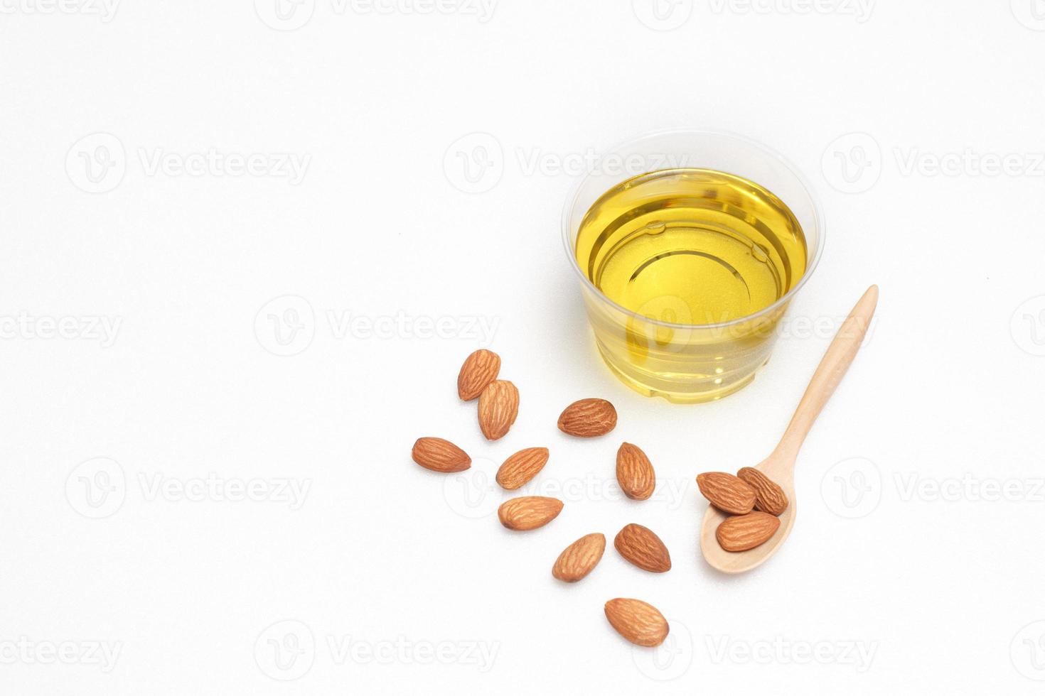 Yellow almond oil in a clear bowl with a wooden spoon next to it and several almonds on a white background photo