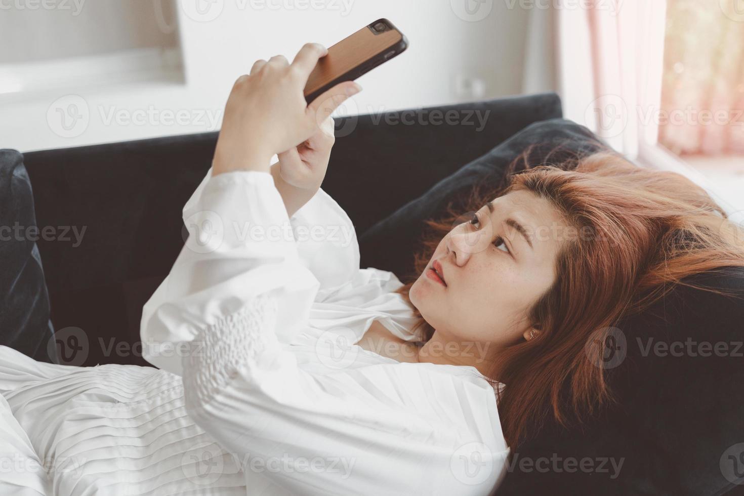 Woman in white dress lying on a sofa in the living room playing mobile phone. Concept of life and relax at home. photo