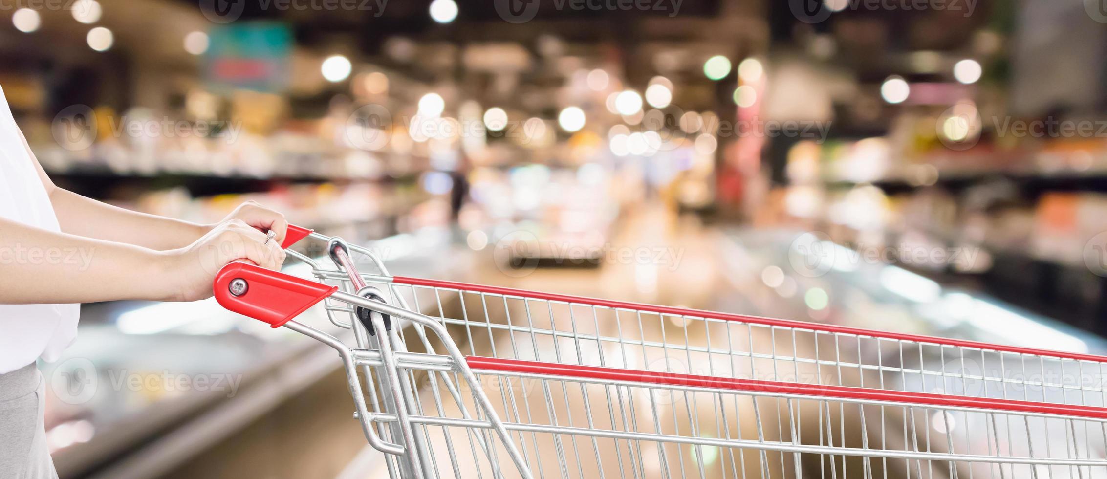 Woman hand hold empty red shopping cart with abstract blur supermarket discount store aisle interior defocused background with bokeh light photo