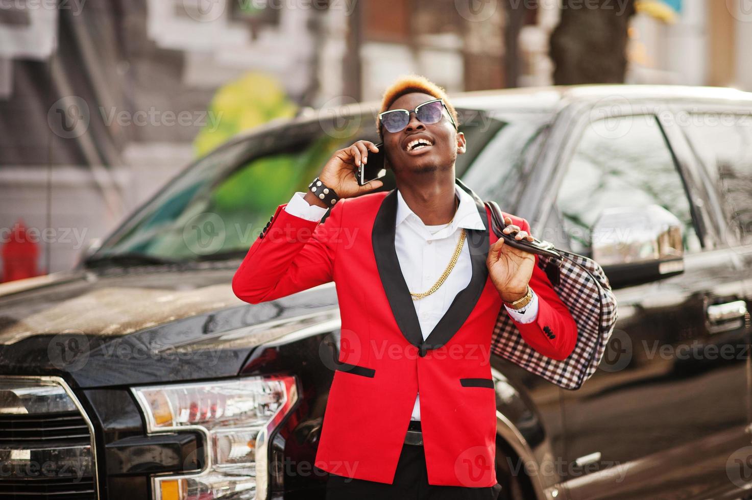 Fashion african american man model at red suit, with highlights hair posed against big suv black car and speaking on mobile phone. photo