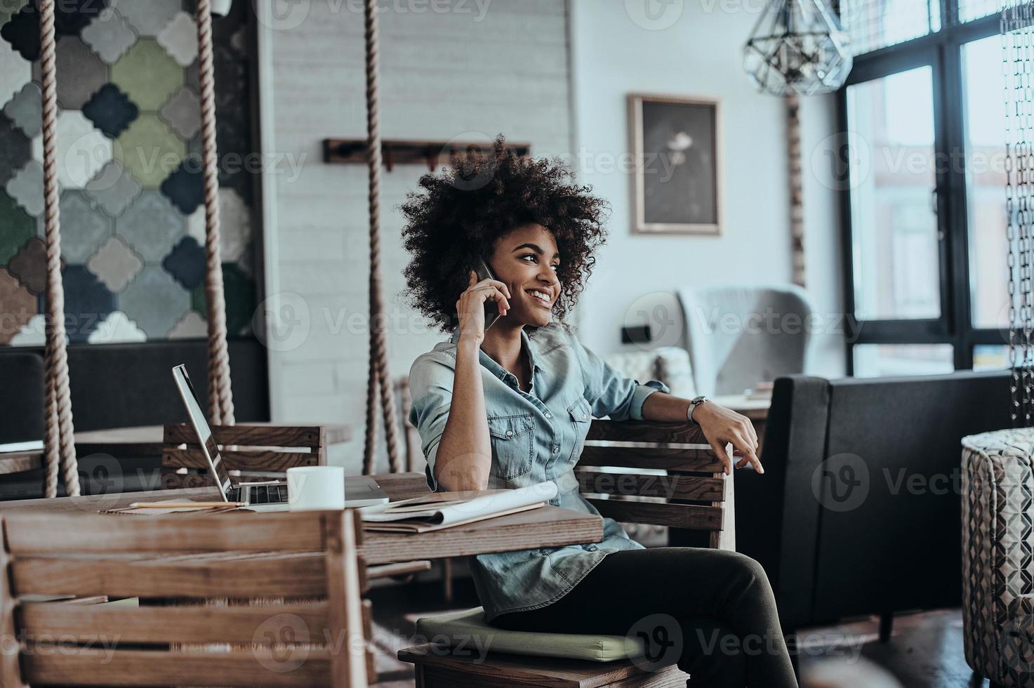 Always in touch with clients. Beautiful young African woman talking on smart phone and smiling while sitting at her working place photo
