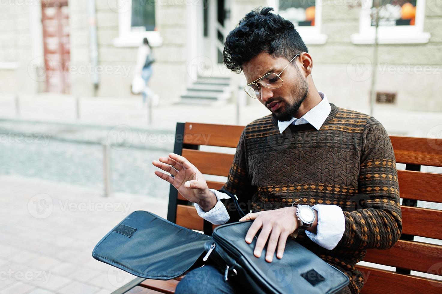 Portrait of young stylish indian man model pose in street, sitting on bench with handbag. photo