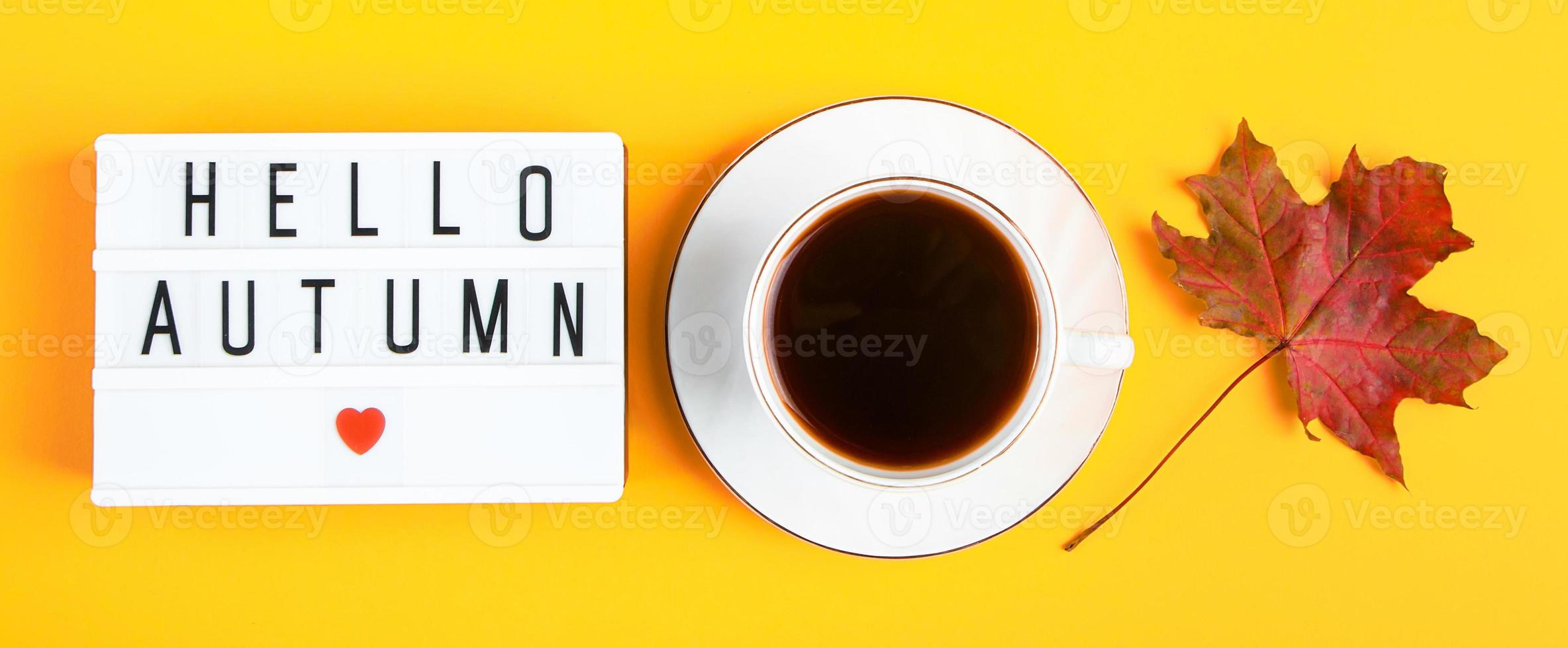 Hello autumn concept. Cup of coffee and sign with inscription on yellow background. Fallen leaves and hot drink. photo