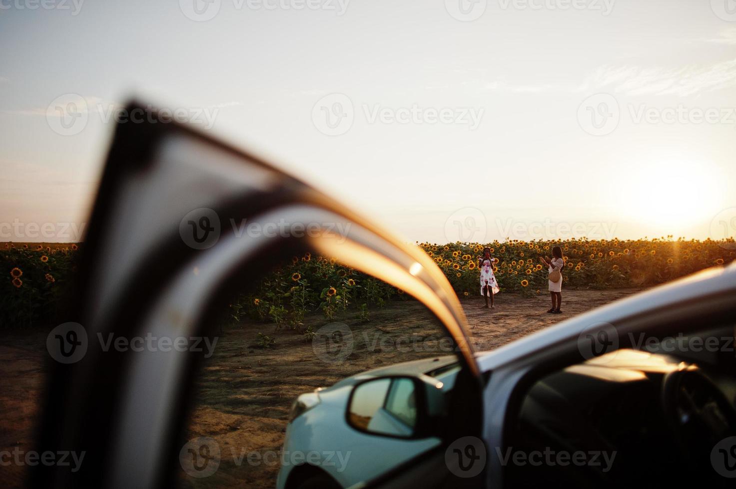 White car against two pretty black womans wear summer dress pose in a sunflower field. photo