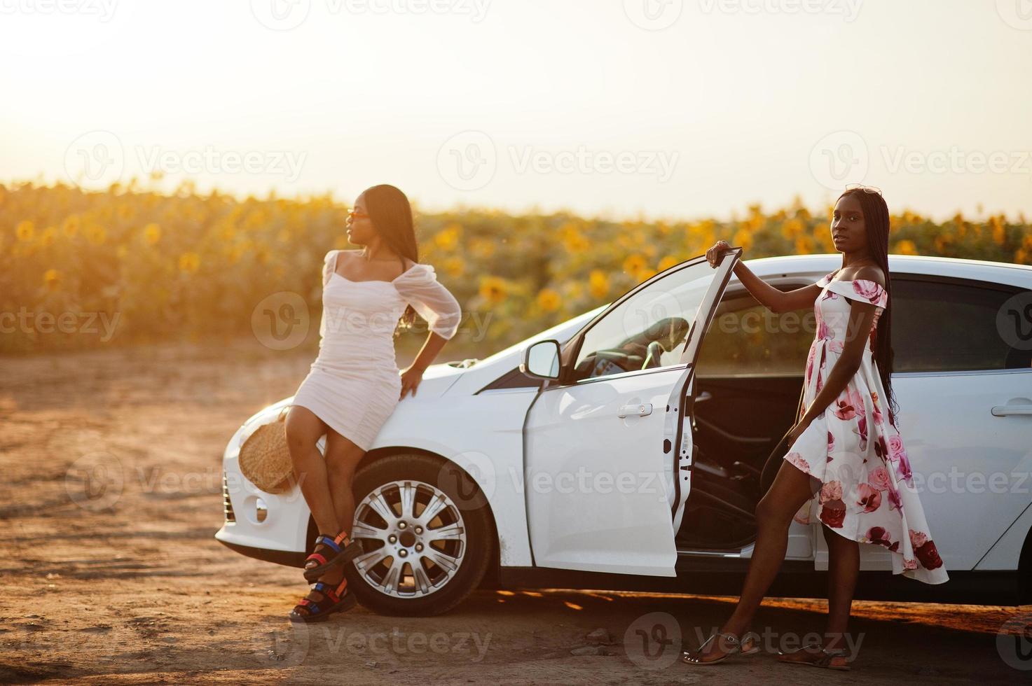 Two pretty young black womans wear summer dress pose in car against a sunflower field. photo