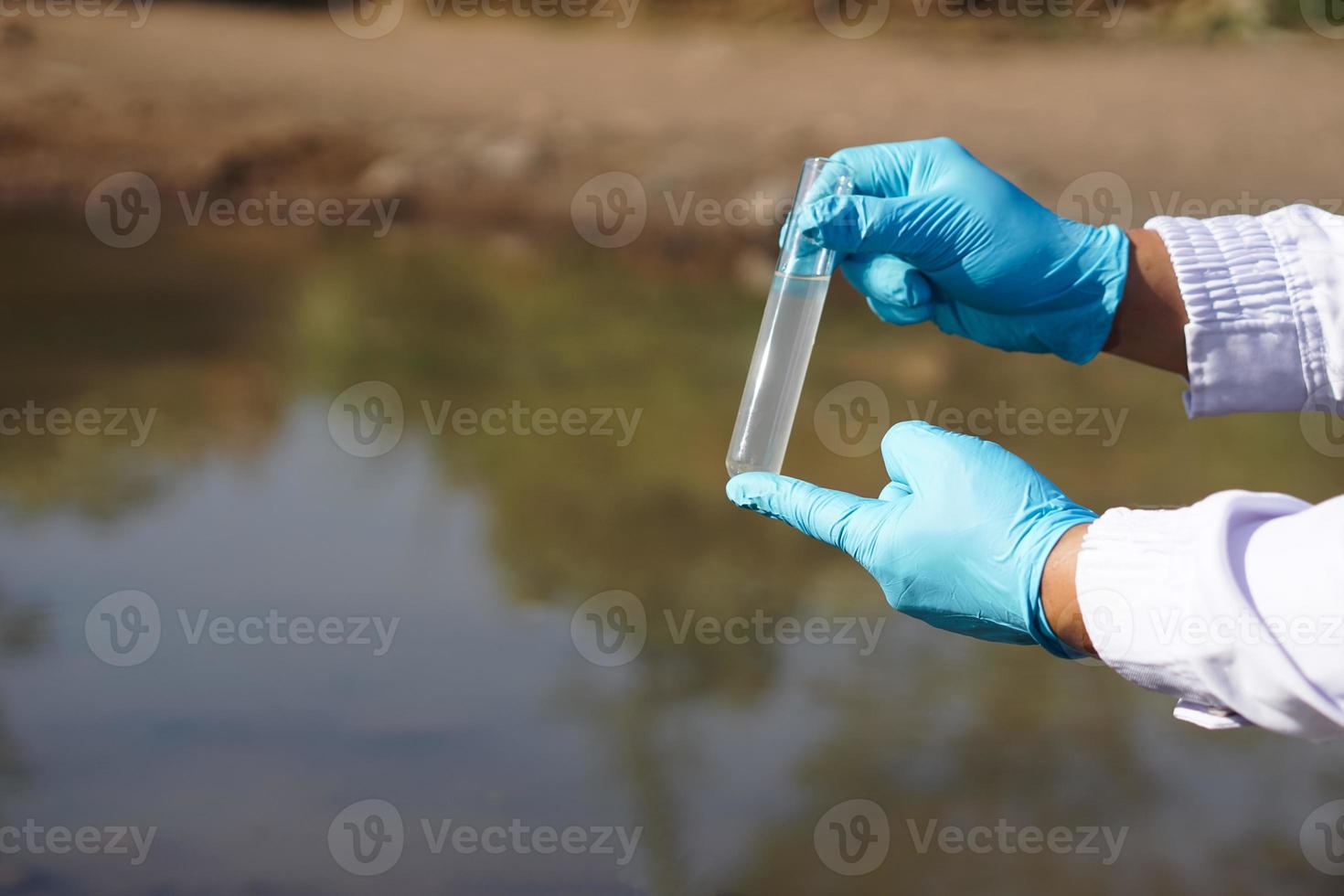 las manos del investigador de primer plano usan guantes azules y sostienen un tubo de vidrio de prueba que contiene agua de muestra del lago. concepto, explorar, inspeccionar la calidad del agua de fuente natural. foto