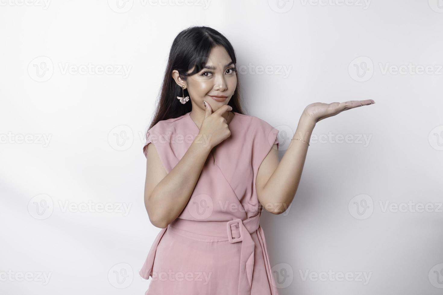 A thoughtful young woman dressed in pink and holding her chin while pointing copy space beside her, isolated by white background photo