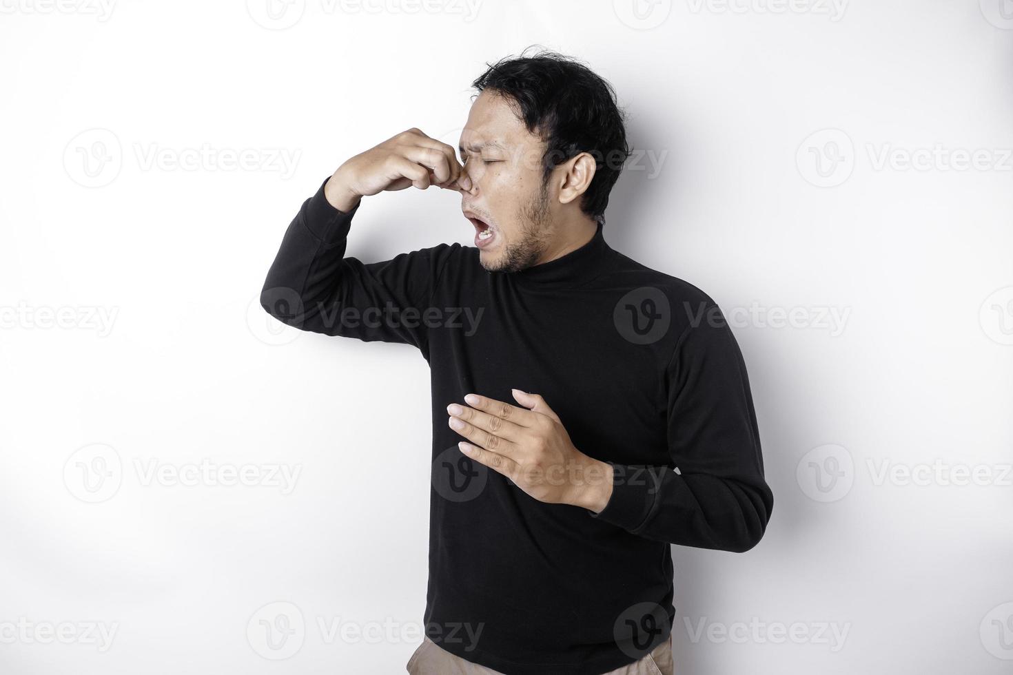 Young Asian man wearing t-shirt standing over isolated white background smelling something stinky and disgusting, intolerable smell, holding breath with fingers on nose. Bad smells concept. photo