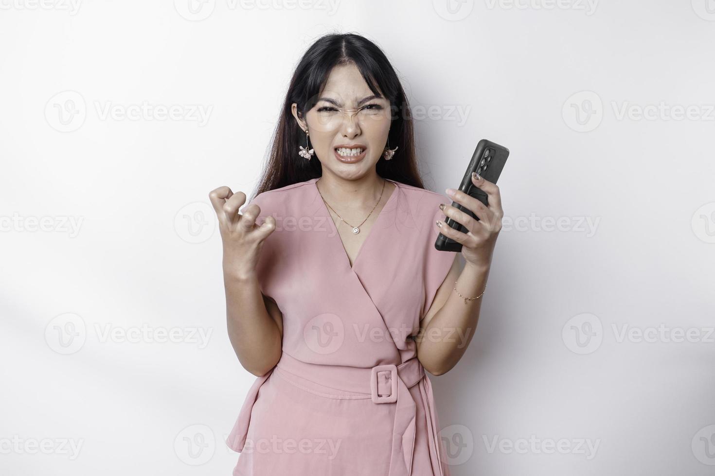 un insatisfecho joven asiático mujer vestido en rosa, mira descontento con irritado cara expresiones participación su teléfono foto
