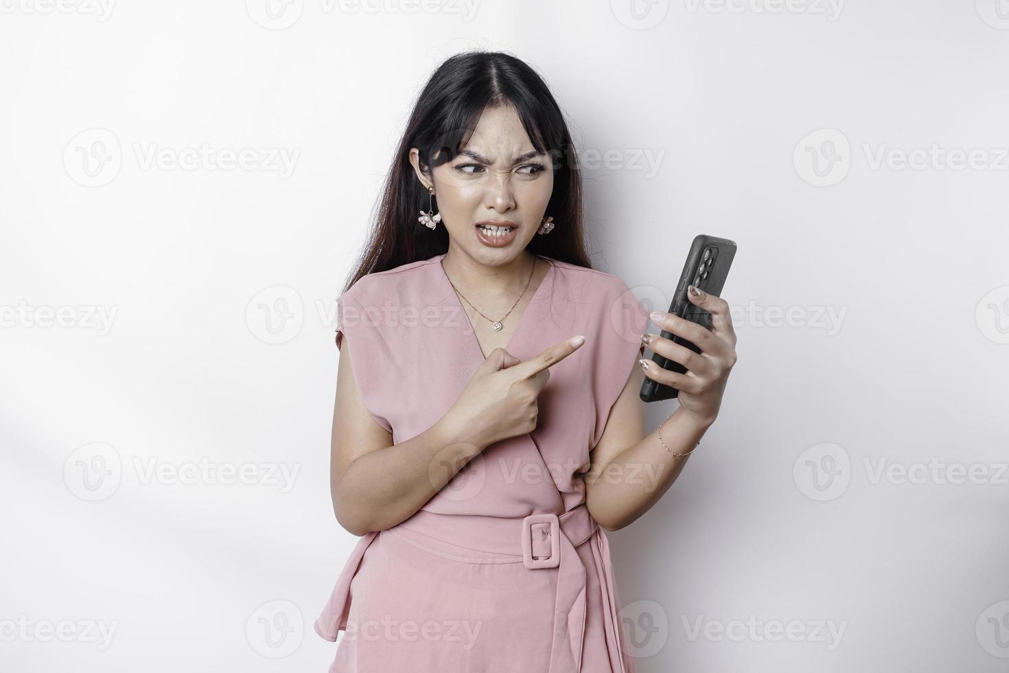 A dissatisfied young Asian woman dressed in pink, looks disgruntled with irritated face expressions holding her phone photo