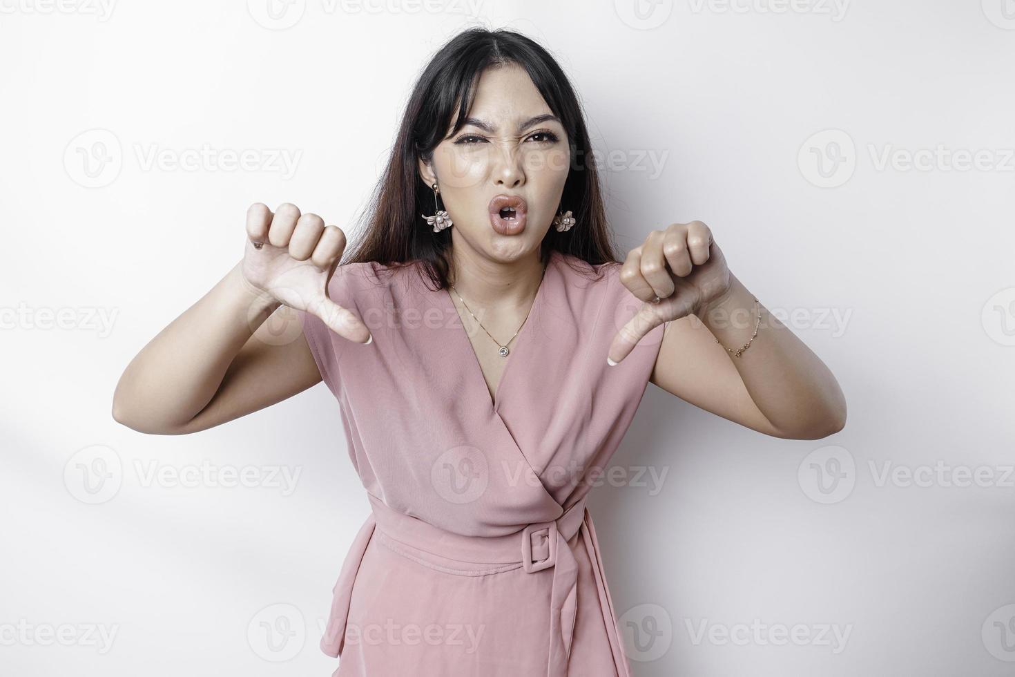 Disappointed Asian woman wearing pink blouse gives thumbs down hand gesture of disapproval, isolated by white background photo