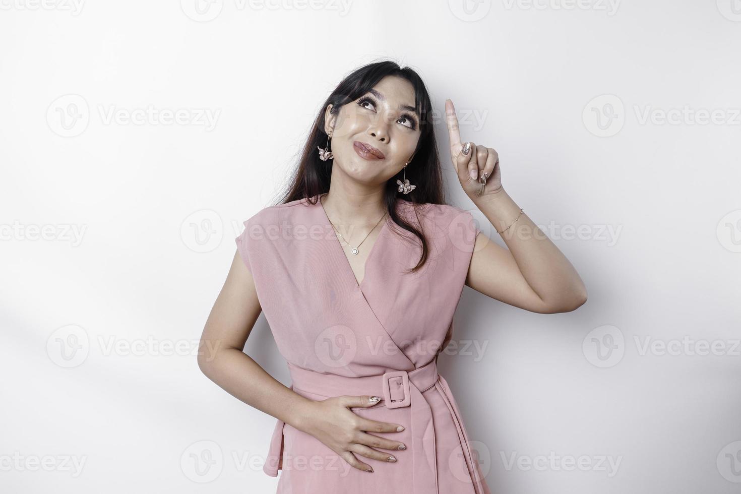 Starving Asian woman dressed in pink, pointing at the copy space on top of her, isolated by white background photo