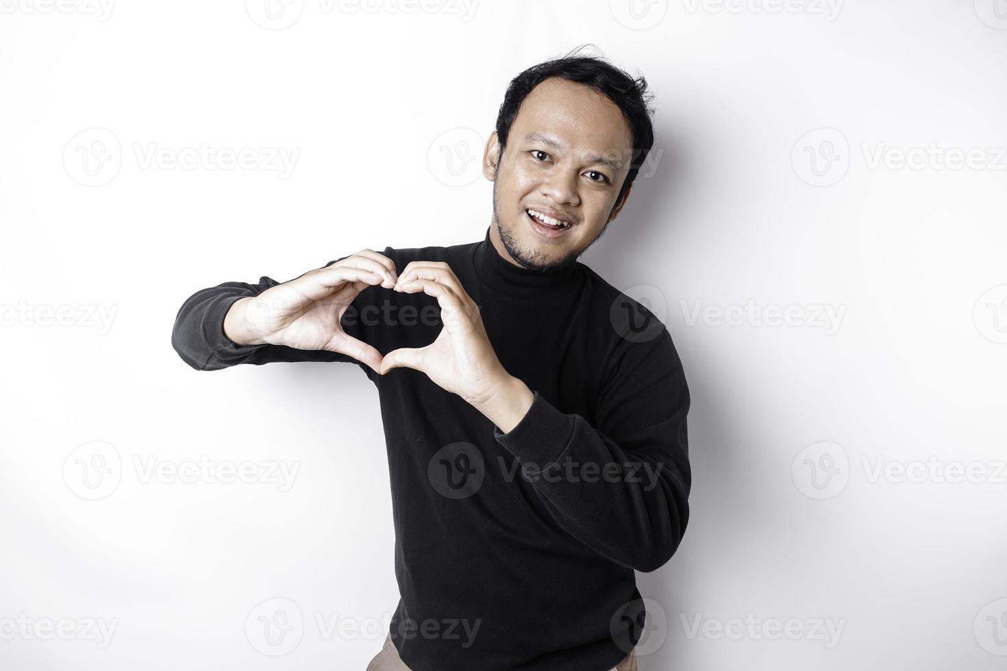 un contento joven asiático hombre vistiendo un negro camisa siente romántico formas corazón gesto expresando oferta sentimientos foto