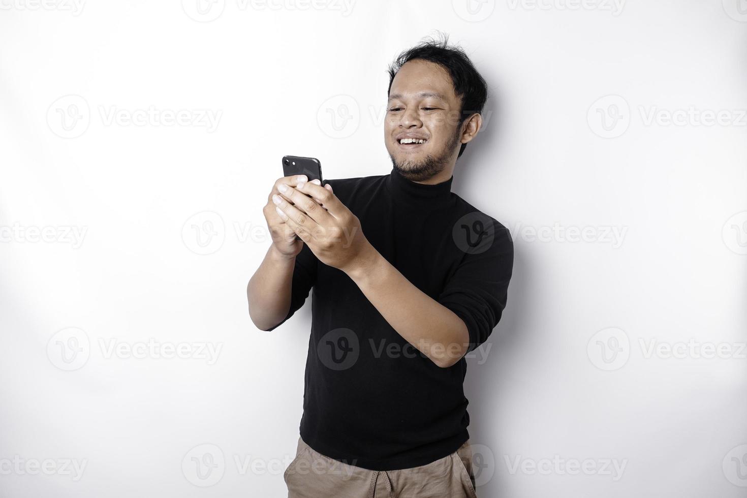 emocionado asiático hombre vistiendo negro camisa sonriente mientras participación su teléfono, aislado por blanco antecedentes foto