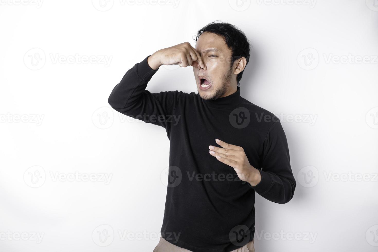 Young Asian man wearing t-shirt standing over isolated white background smelling something stinky and disgusting, intolerable smell, holding breath with fingers on nose. Bad smells concept. photo