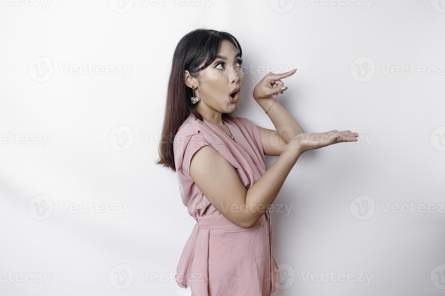 Shocked Asian woman dressed in pink, pointing at the copy space on beside her, isolated by white background photo