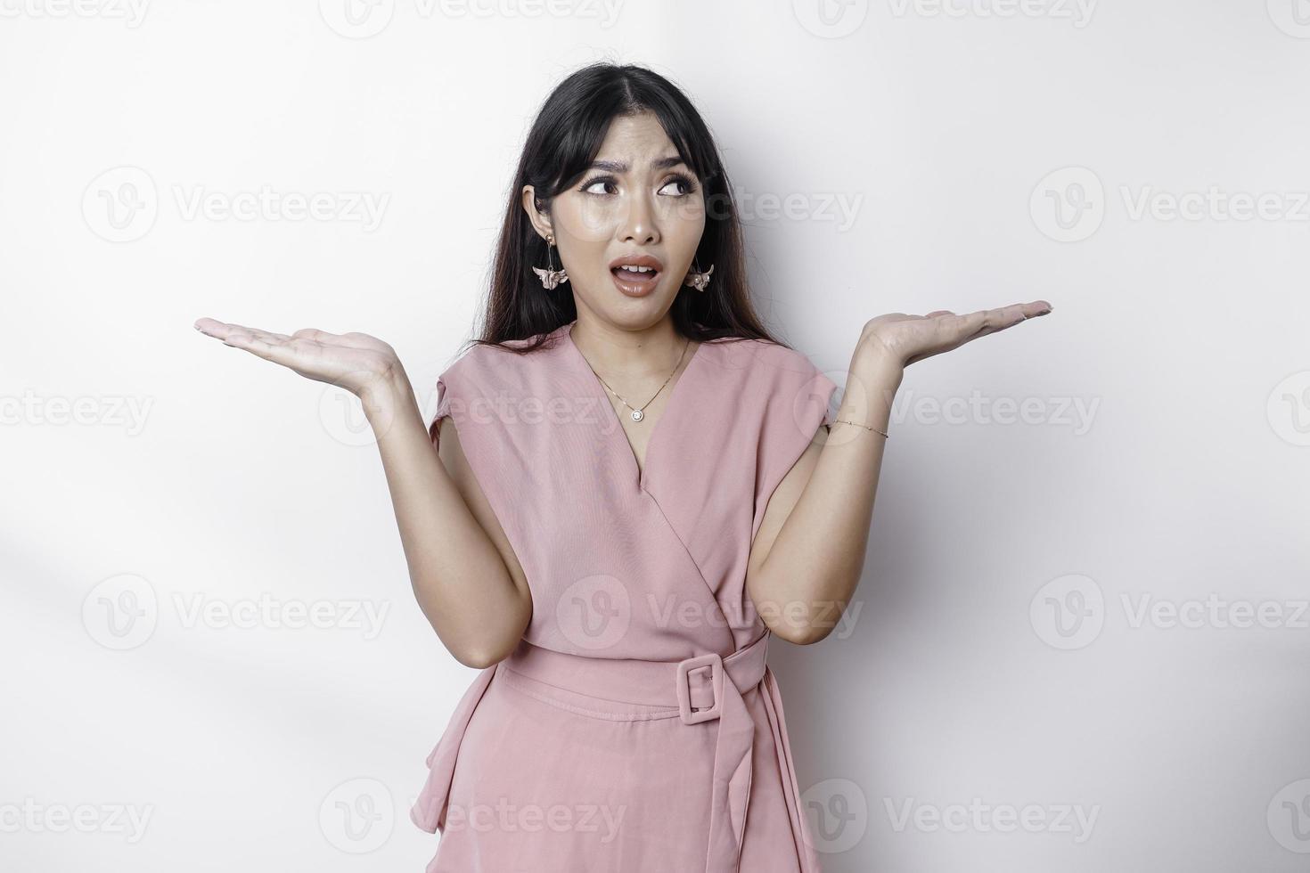 un pensativo joven asiático mujer es vistiendo rosado blusa participación su teléfono y mira confundido, aislado por blanco antecedentes foto