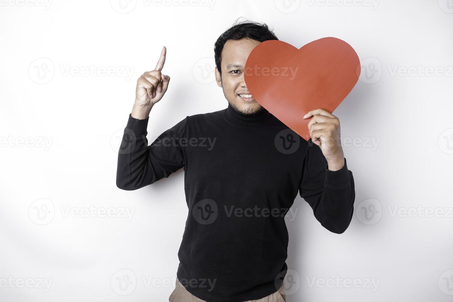 Portrait of a smiling Asian man holding a big red heart symbol pointing up at copy space isolated over white background photo