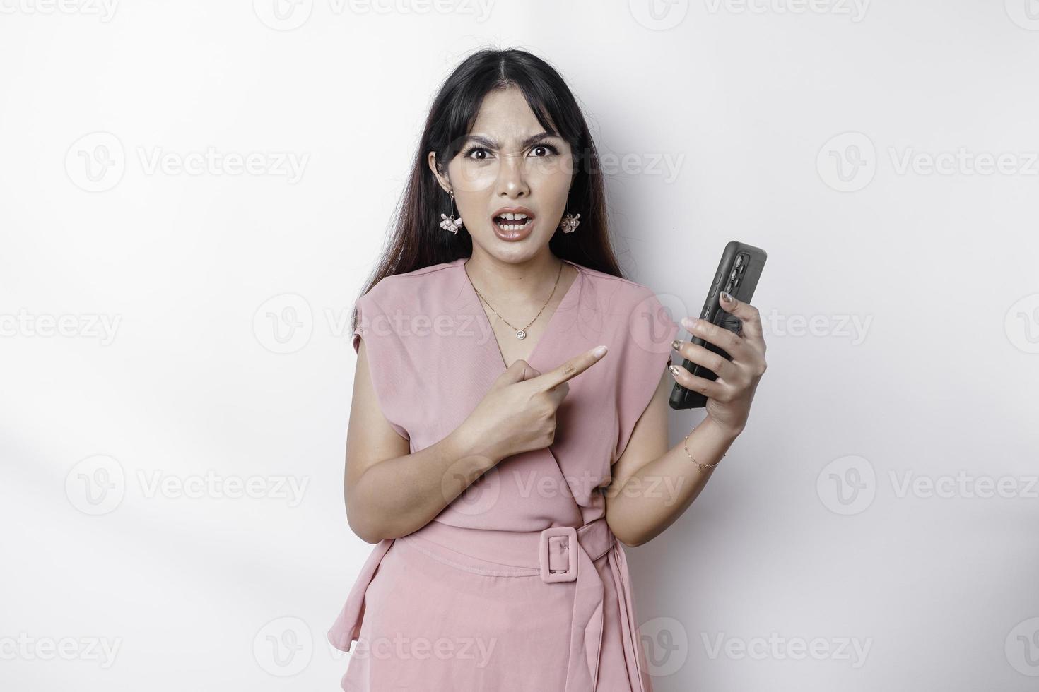 A dissatisfied young Asian woman dressed in pink, looks disgruntled with irritated face expressions holding her phone photo