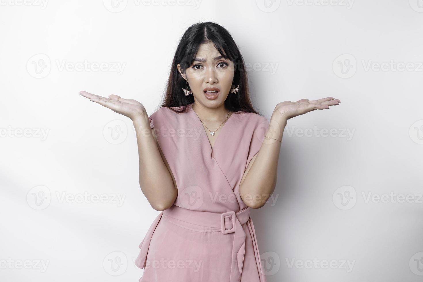 un pensativo joven asiático mujer es vistiendo rosado blusa participación su teléfono y mira confundido, aislado por blanco antecedentes foto