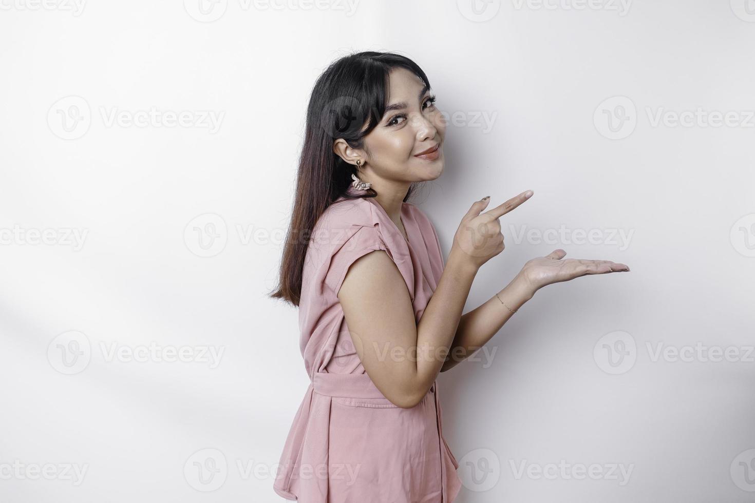 Excited Asian woman dressed in pink, pointing at the copy space beside her, isolated by white background photo
