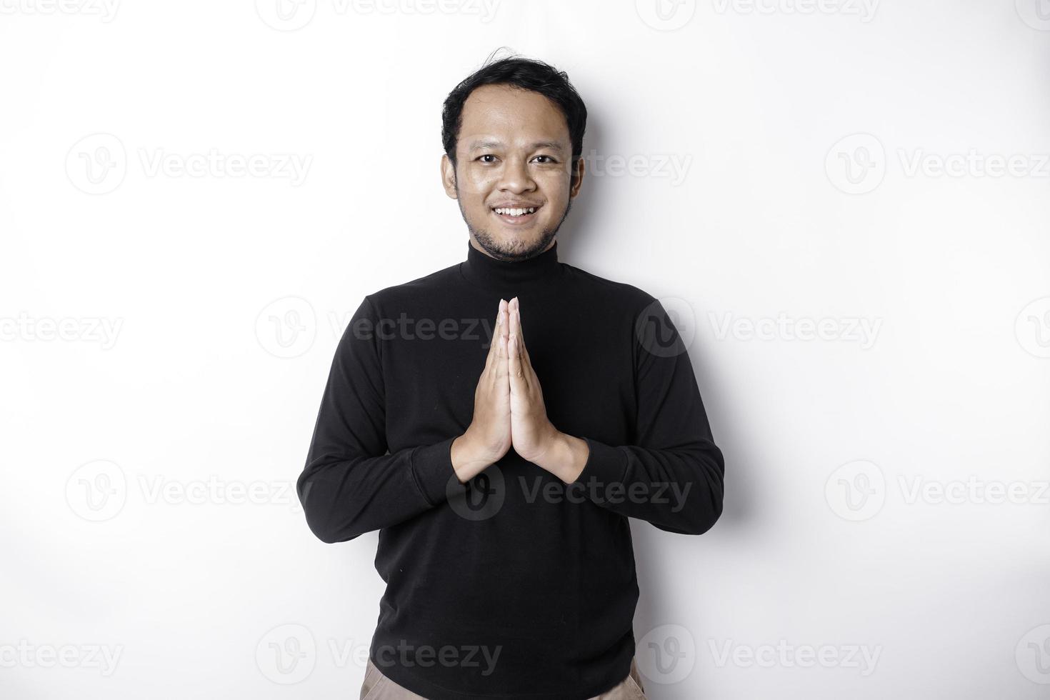 Smiling young Asian man wearing black shirt, gesturing traditional greeting isolated over white background photo