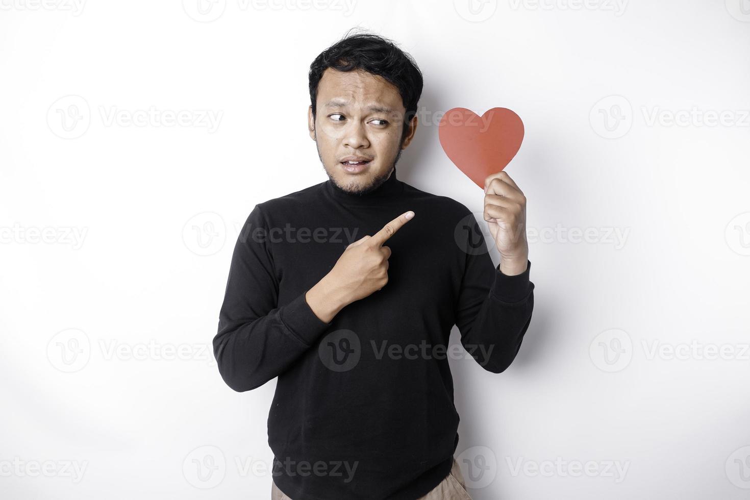 A portrait of an Asian man wearing a black shirt looks so confused while holding a red heart-shaped paper, isolated by a white background photo