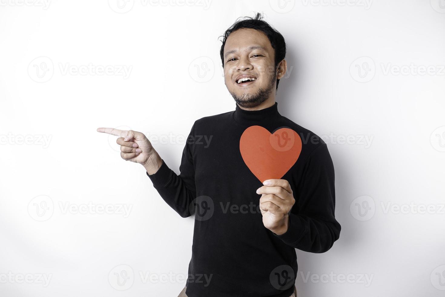 Excited Asian man wearing black shirt, pointing at the copy space beside him while holding a big red heart-shaped paper, isolated by white background photo