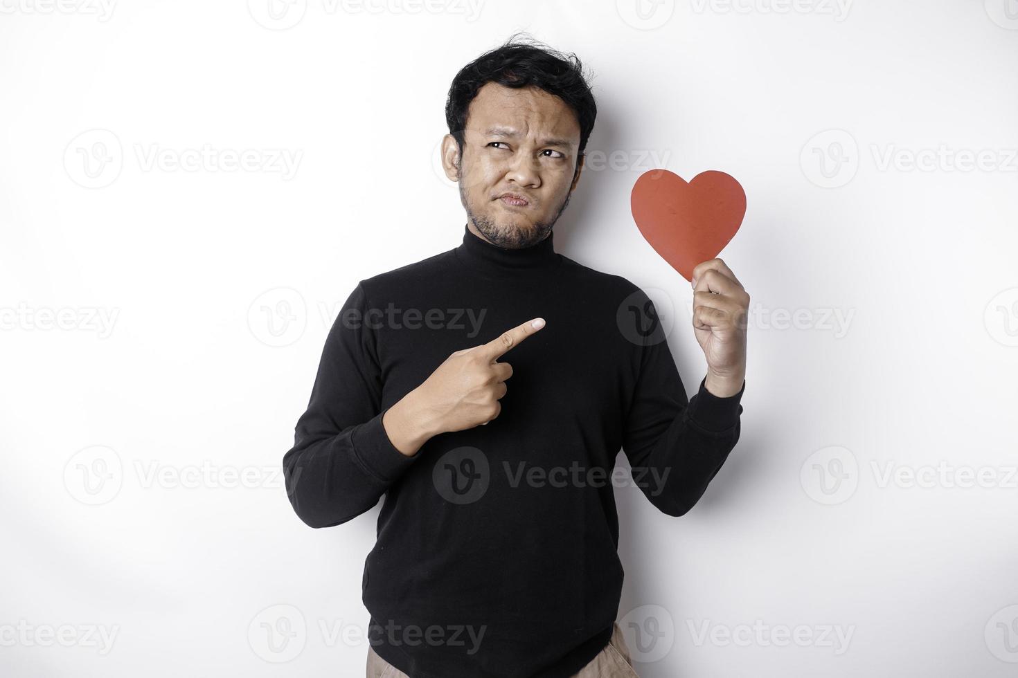 A portrait of an Asian man wearing a black shirt looks so confused while holding a red heart-shaped paper, isolated by a white background photo