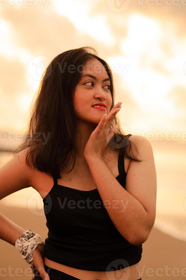 un asiático Adolescente niña en un negro camisa con un sonriente expresión soportes en frente de el arena o con el olas estrellarse mientras disfrutando el ver foto