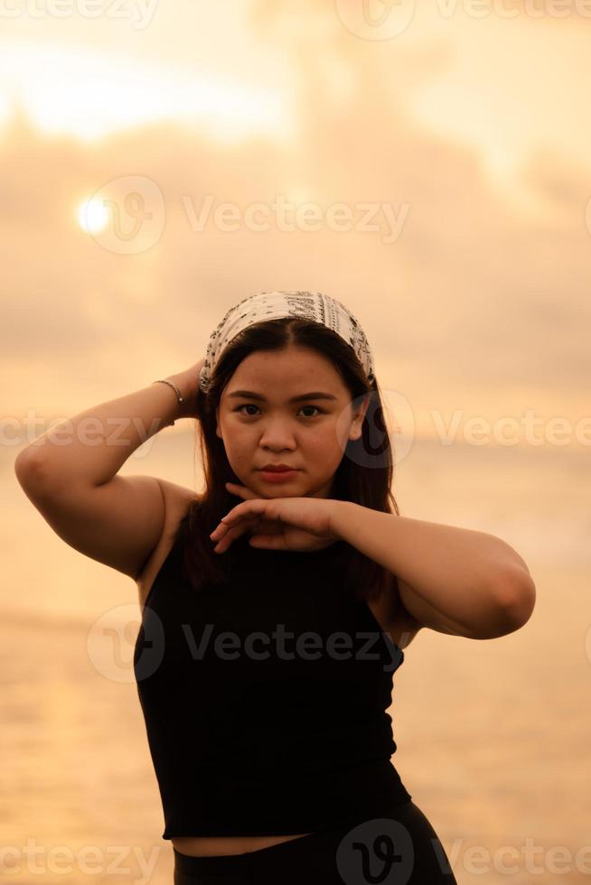 a glamorous Asian woman in black clothes and a white bandana standing in front of the waves on the beach photo