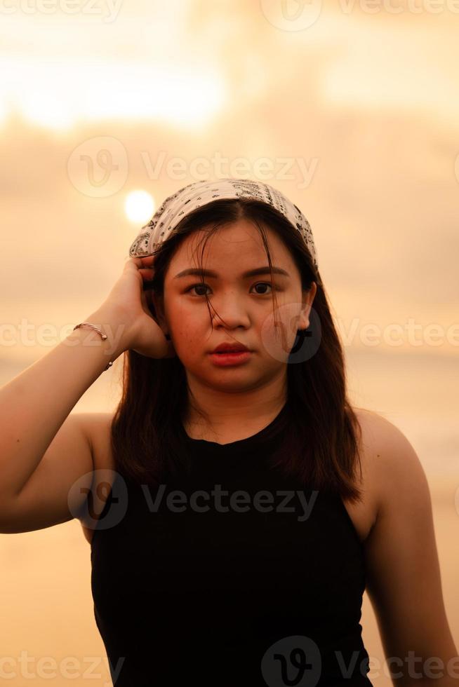 an Asian teenager wearing a white bandana and black shirt with a flat expression while posing with her hair on the beach photo