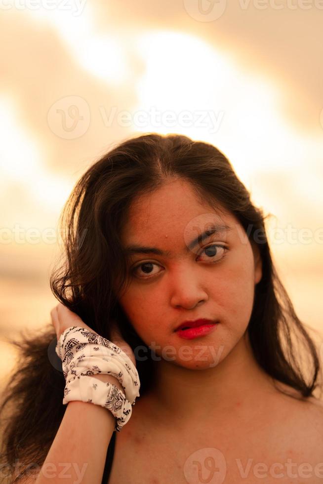 An Asian woman in a black shirt with an expression that looks annoyed is standing near the waves on the beach in a holiday photo