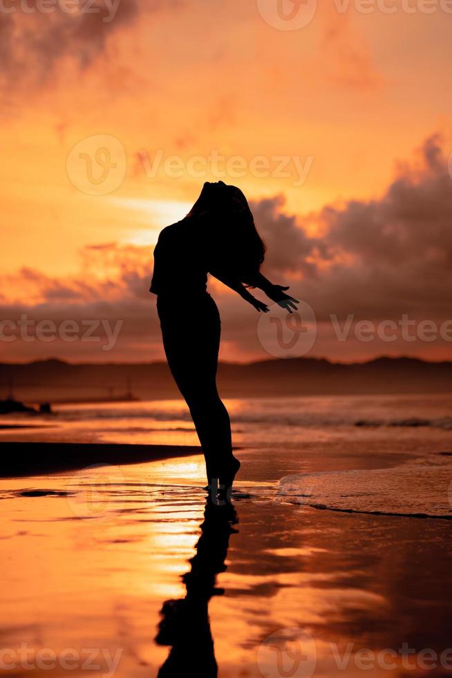 an Asian woman in silhouette is doing a very beautiful dance on the beach with the waves crashing photo