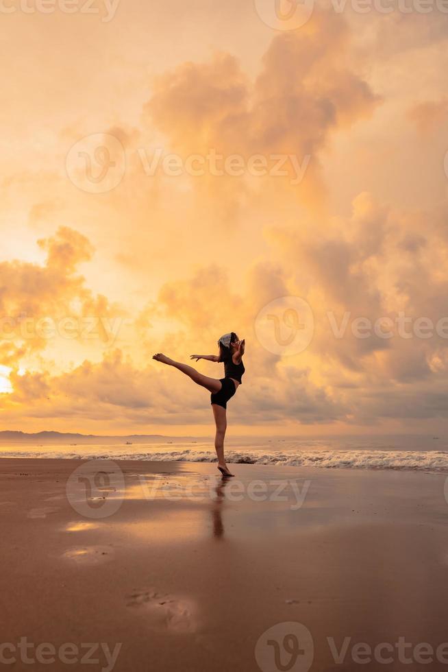 A ballerina in a black dress is practicing ballet moves on the beach with very flexible movements with a view of the clouds behind photo