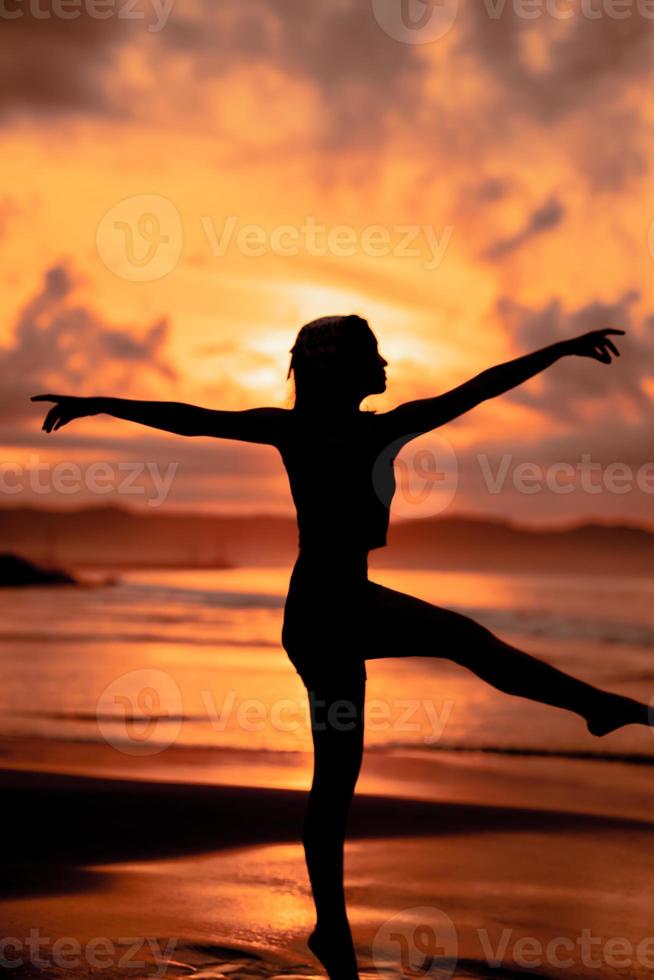 A ballerina with a silhouette shape performs ballet movements very flexibly on the beach with the waves crashing photo