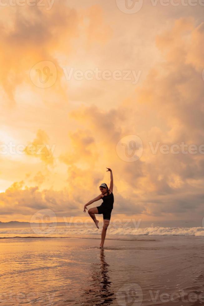 An Asian woman in a black dress performs ballet movements beautifully on the beach with waves and clouds behind her photo
