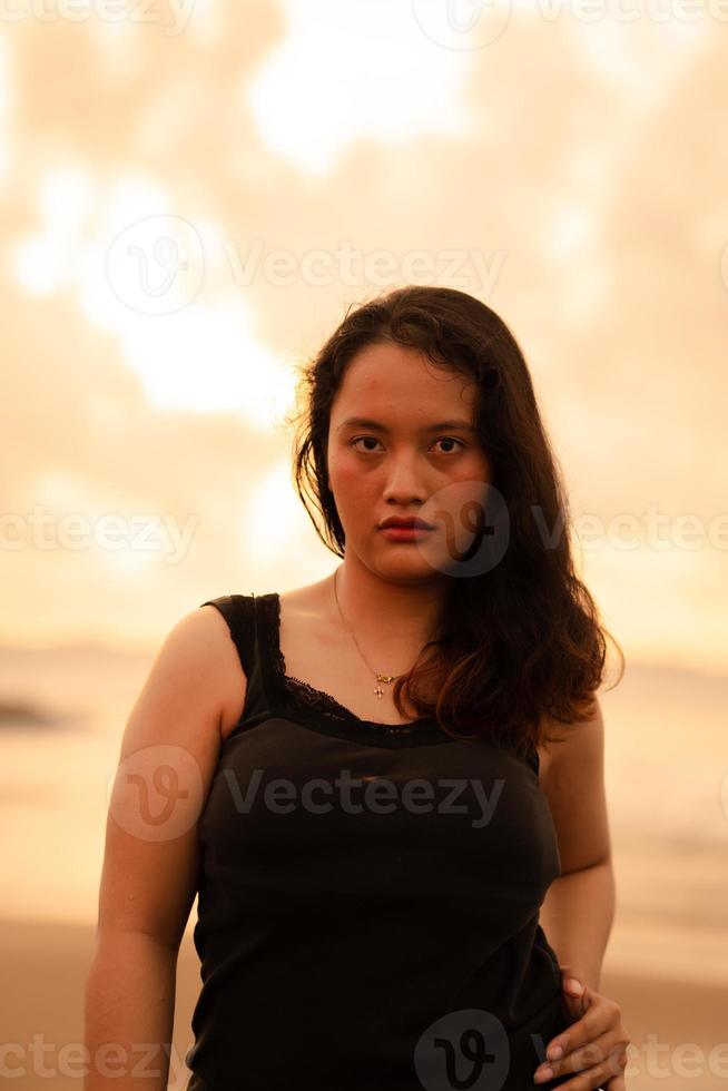 retrato imagen de un asiático mujer con negro pelo y un enojado expresión en pie en el playa en su negro ropa foto