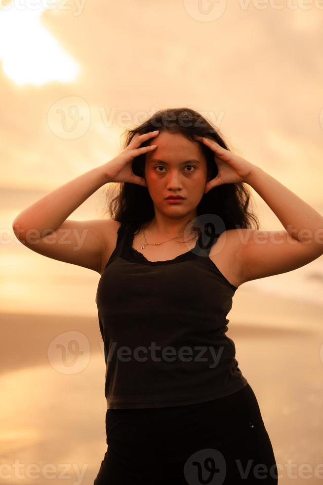 un asiático mujer poses con un negrita y sexy expresión mientras vistiendo negro ropa en frente de el playa foto