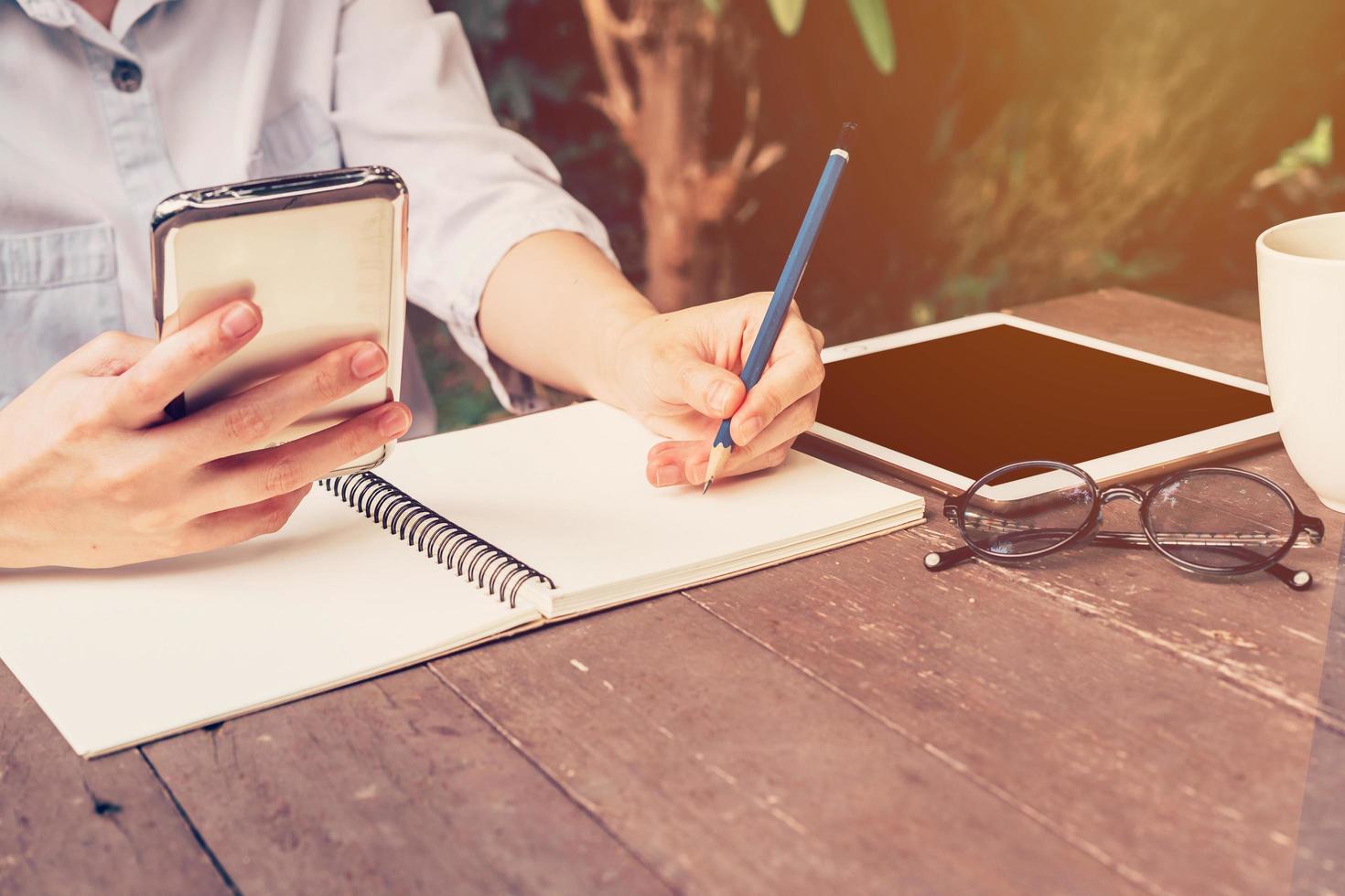 asian woman hand holding phone and pencil writing notebook in coffee shop with vintage toned photo