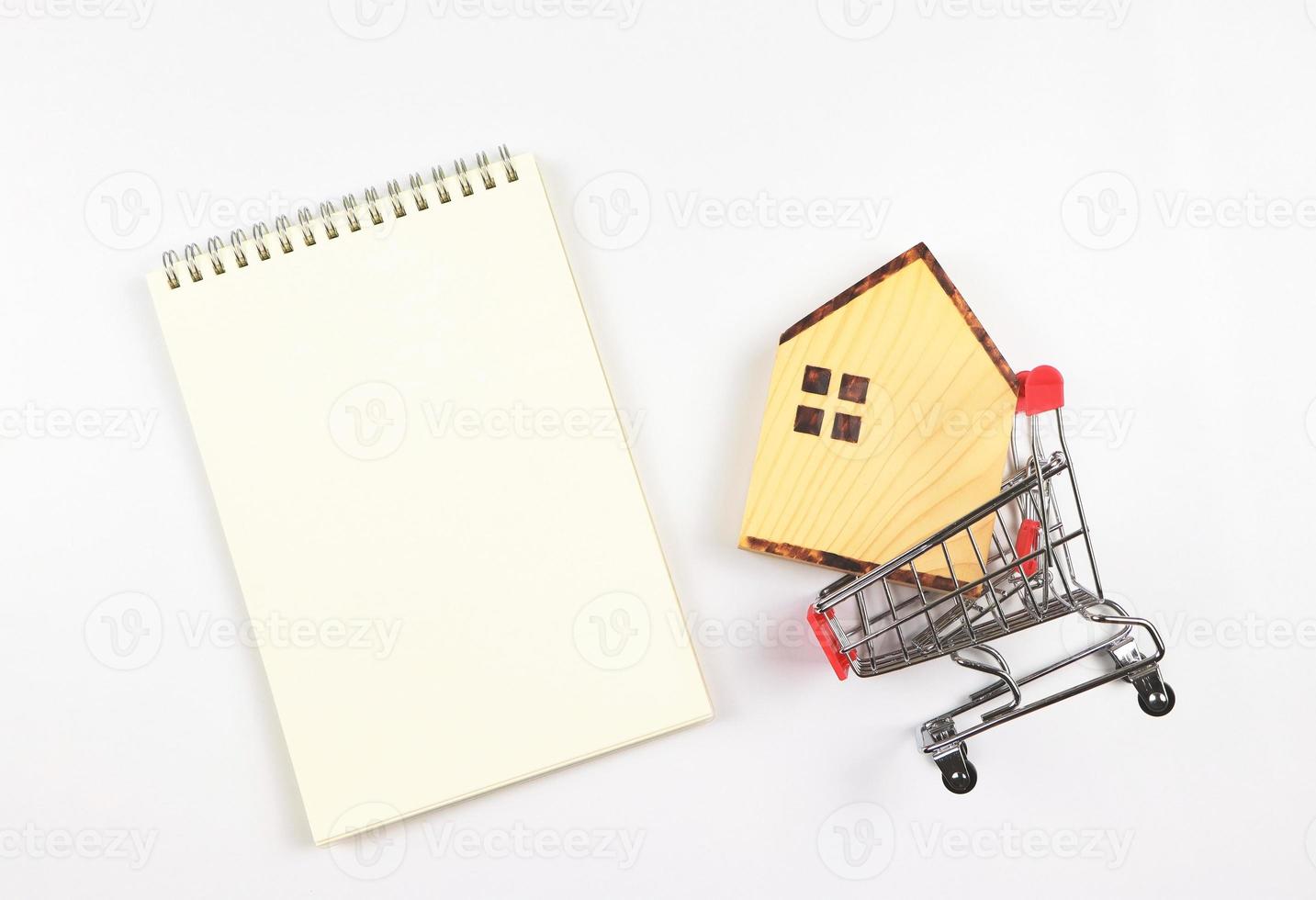 flat layout of wooden house model in shopping trolley or shopping cart with blank page opened notebook on white background, home purchase concept. photo